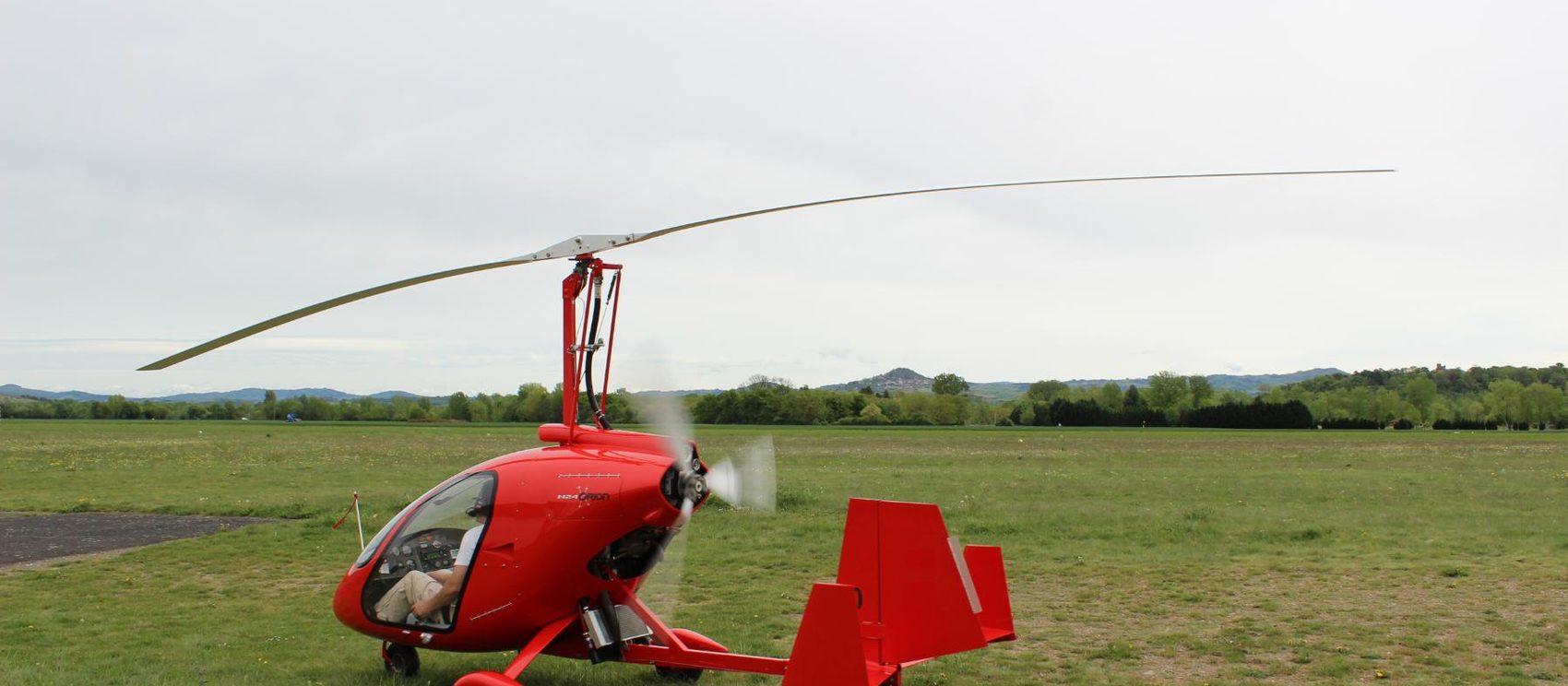 Red open-cockpit gyroplane on a grass airfield with the rotor turning