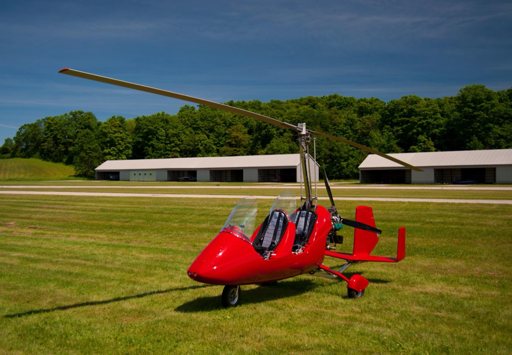 A red open tandem gyrocopter of the patrol-style type parked by hangars.
