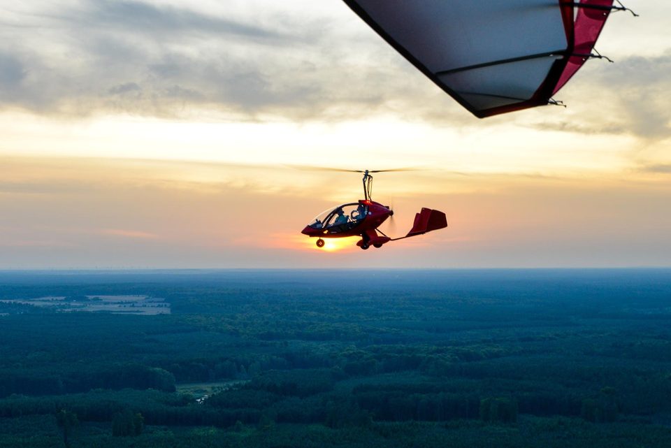 A red gyrocopter cruising low over forest in evening light.