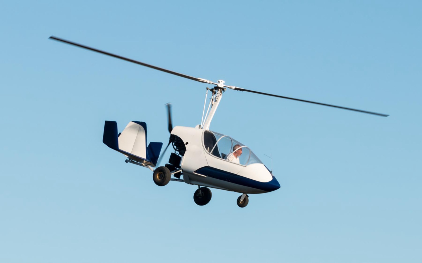 A white and blue gyrocopter in cruise flight against a clear sky.