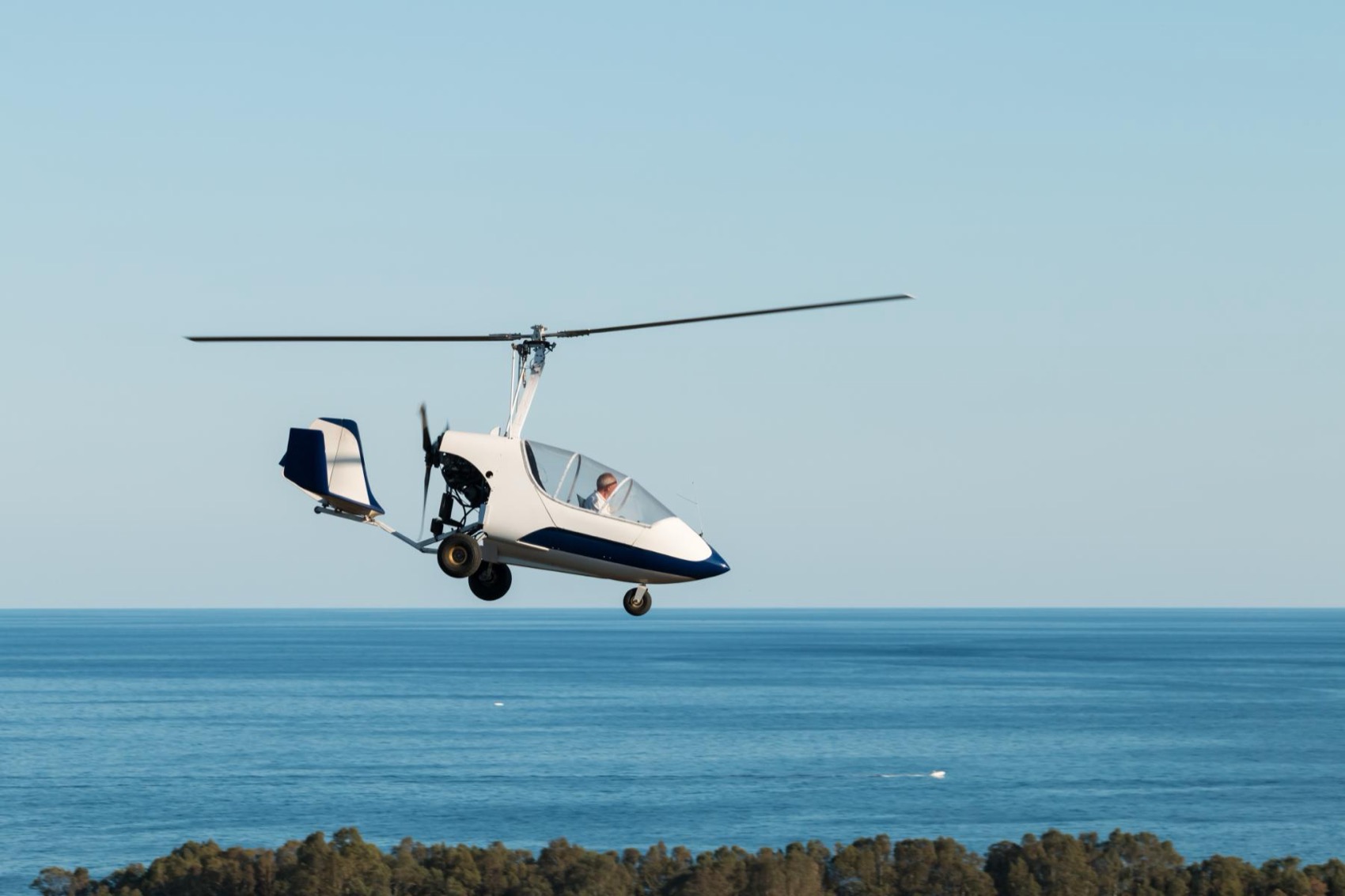 A white gyrocopter gliding low over the sea, rotor autorotating.