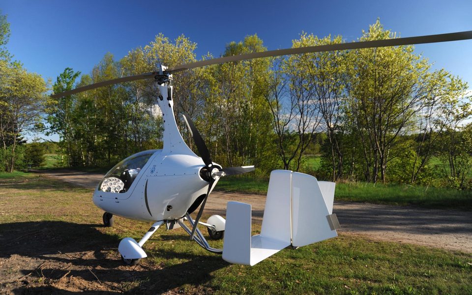 A white enclosed gyrocopter parked on a woodland trail with its rotor at rest.