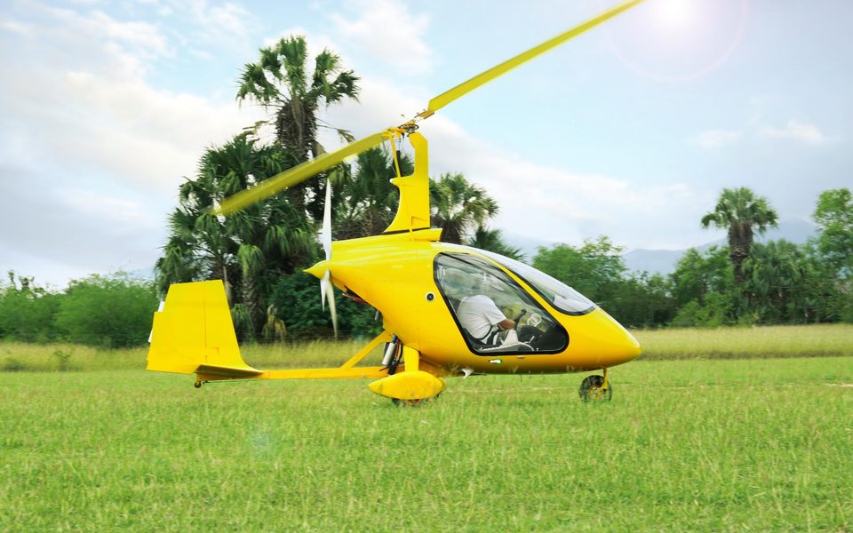 A yellow gyrocopter on grass with its rotor turning during pre-rotation before takeoff.